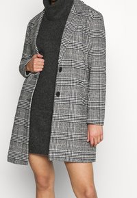 Mujer vistiendo un vestido de punto negro con cuello alto debajo de un abrigo largo de cuadros gris y negro con botones negros.