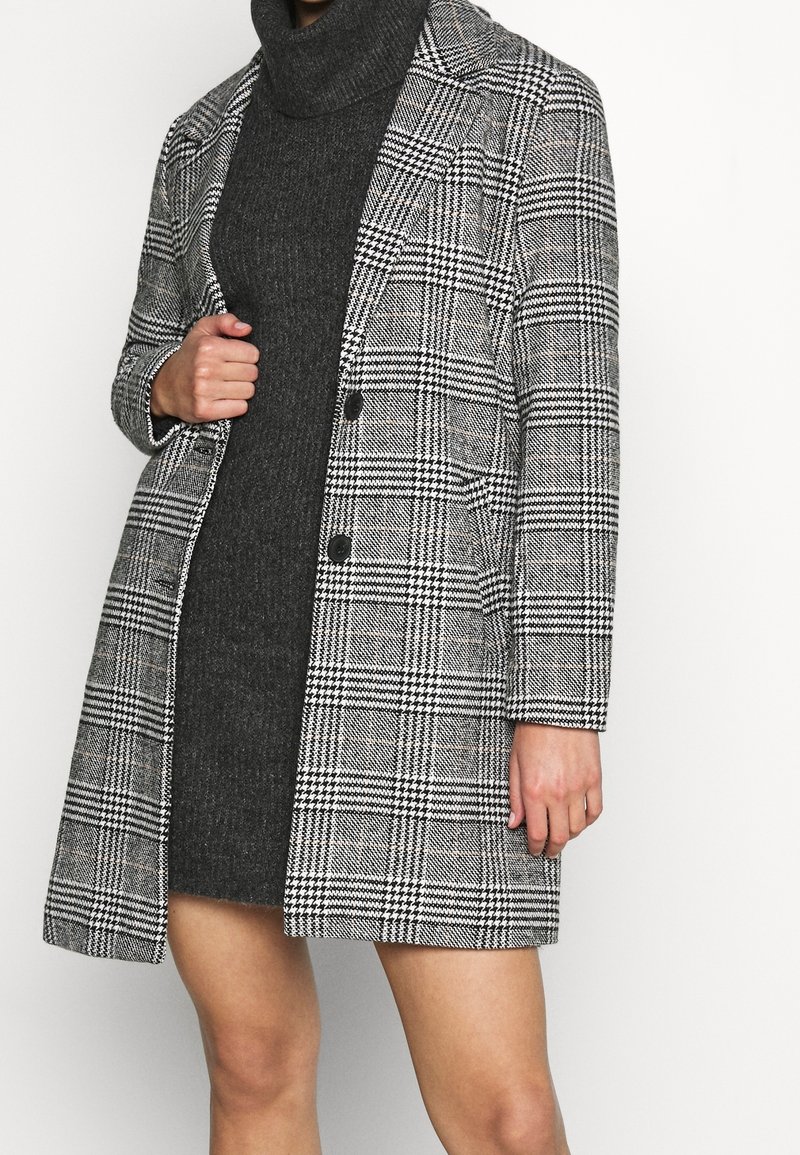 Mujer vistiendo un vestido de punto negro con cuello alto debajo de un abrigo largo de cuadros gris y negro con botones negros.