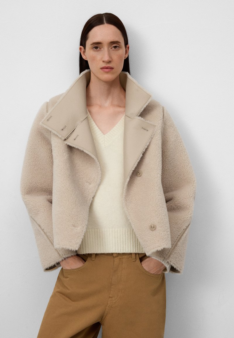 Beżowa kurtka z owczej wełny z szerokim kołnierzem, podwójnymi guzikami i miękką teksturą, noszona na jasnym kremowym swetrze i brązowych spodniach.