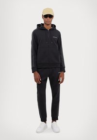 Black zip-up hoodie with white stripes, paired with matching black sweatpants. White sneakers complete the look, featuring a subtle logo.