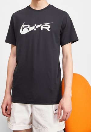 Osoba nosząca czarną koszulkę z białym "AIR" i logo typu swoosh oraz białe spodenki, stojąca obok pomarańczowego krzesła na jasnym tle.