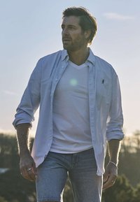 Hombre con mangas remangadas, lleva una camisa de rayas sobre una camiseta blanca y pantalones cortos de mezclilla, de pie al aire libre con árboles y cielo de fondo.