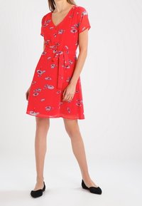 Červené šaty s krátkým rukávem a výstřihem do V, květinovým vzorem, detailem knoflíků a šňůrkou v pase. Lehká tkanina s splývavým designem.