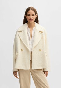 Dubbelt knäppt cremfärgat ullblandat kappa med stora, skårade slag, strukturerad yta och knappdetaljer, i kombination med en vit blus och beige byxor.