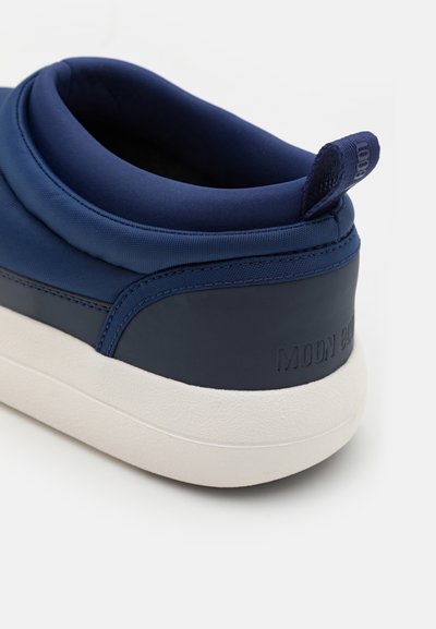 Tumši zilas krāsas slip-on kedas ar strukturētu augšdaļu, mīkstinātu papēža daļu, kontrastējošu baltu zoli un iegravētu "MOON BOOT" logo uz papēža.
