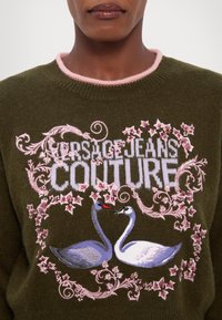Maglione verde lavorato a maglia con colletto a coste rosa, caratterizzato da un'accurata ricamata di cigni e motivi floreali, etichettato "Versace Jeans Couture."