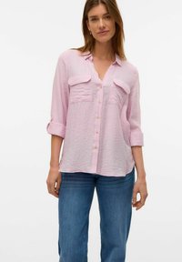 Camisa de botones de un tono rosa claro con un acabado texturizado, que cuenta con dos bolsillos en el pecho y mangas enrolladas de tres cuartos. Combinada con unos vaqueros de mezclilla azul.