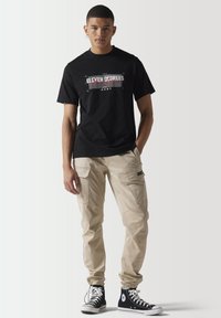 Camiseta negra con estampado gráfico, pantalones cargo beige con bolsillos y zapatillas altas negras con acentos blancos.