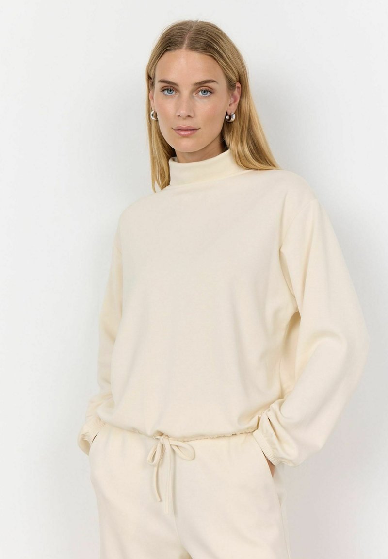 Krémový sweatshirt s vysokým golierom vyrobený z mäkkej tkaniny, s dlhými rukávmi a voľným strihom, doplnený zodpovedajúcimi nohavicami na šnúrku.