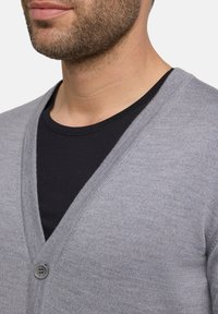 Cardigan grigio con scollatura a V e chiusura a bottoni, indossato sopra una maglietta nera a girocollo. Texture morbida e sottile motivo a maglia visibile.