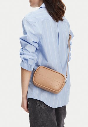 Mujer con camisa de rayas azul claro y pantalones negros, llevando un pequeño bolso beige acolchado de bandolera sobre el hombro.