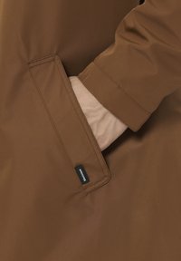 Rjava vodoodporna jakna z gladko teksturo, s stranskim žepom in znamčeno etiketo na šivu žepa.