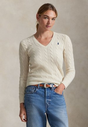 Ung kvinde med lyst hår iført en cremefarvet cable-knit V-hals sweater og blå jeans, stående mod en beige baggrund.