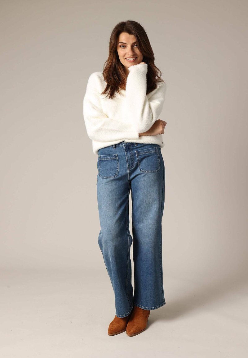 Witte fluffy trui, hooggerande blauwe wijde broek en bruine enkellaarsjes. Eenvoudig ontwerp zonder zichtbare patronen of versieringen.