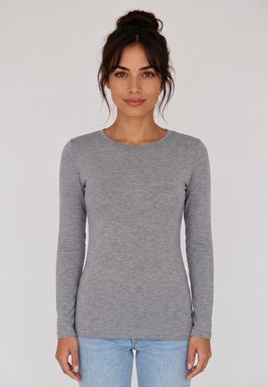 Donna con capelli scuri raccolti in uno chignon, che indossa una maglietta grigia aderente a maniche lunghe e jeans azzurri, in piedi su uno sfondo chiaro e uniforme.