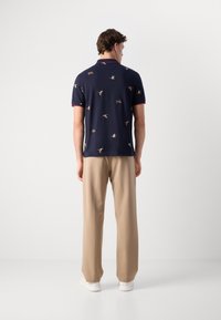 Marine polo skjorte med fargerike fuglemønstre, korte ermer og rød krage, sammen med lyse beige bukser og hvite sko.