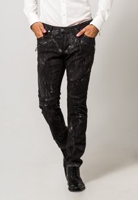 Pantaloni in denim nero con strisce astratte bianche, dotati di tasche con zip, vestibilità slim e polsini alla caviglia. Indossati con una camicia bianca a maniche lunghe.