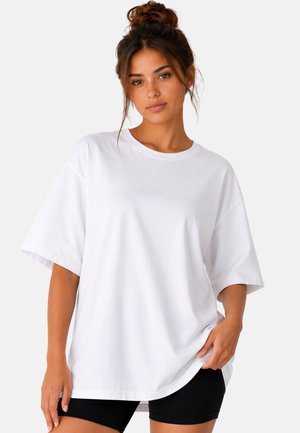 Jeune femme aux cheveux bruns en chignon, portant un t-shirt blanc ample et un short noir, debout devant un fond clair uni.