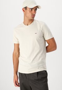 Tricou din bumbac crema, cu mânecă scurtă, cu decolteu rotund și un logo mic care are accente roșii, albe și albastre pe piept.