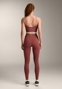 Donna con capelli ricci rossi, indossa leggings color malva a vita alta e un reggiseno sportivo coordinato con cinghie, vista di schiena su uno sfondo neutro.
