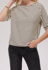 Gestreifter Pullover in Creme und Schwarz, mit ribbeltem Halsausschnitt und kurzen Ärmeln, mit lockerer Passform und strukturiertem Stoff. Getragen mit dunklen Jeans.