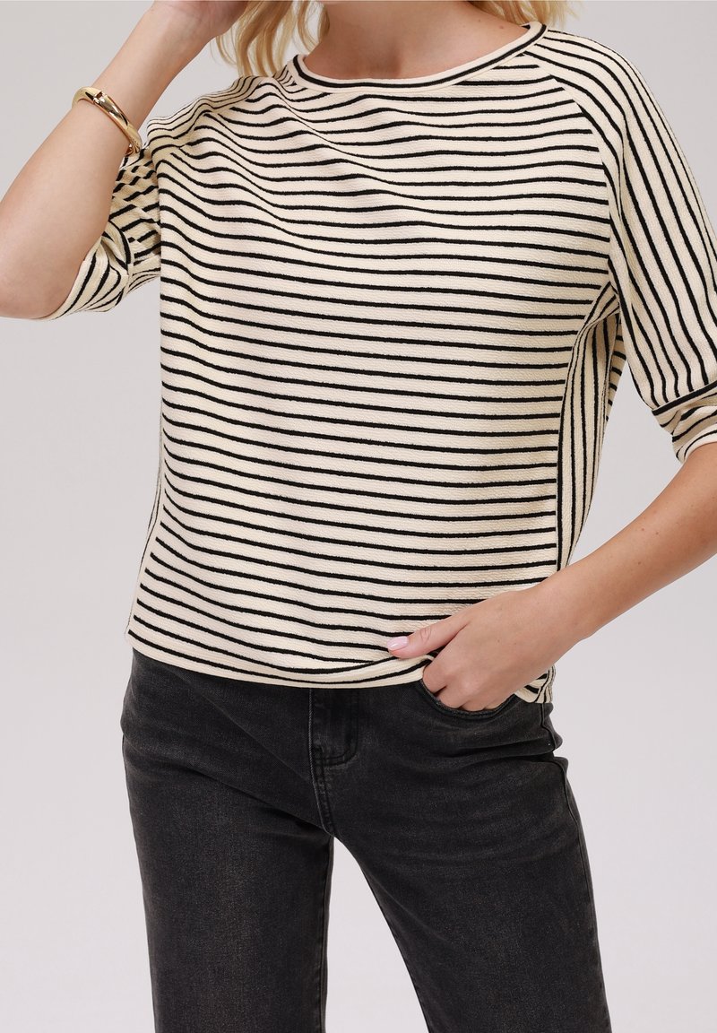 Gestreifter Pullover in Creme und Schwarz, mit ribbeltem Halsausschnitt und kurzen Ärmeln, mit lockerer Passform und strukturiertem Stoff. Getragen mit dunklen Jeans.