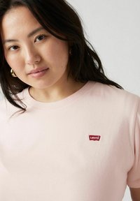 Camiseta de algodón rosa claro con cuello redondo y un pequeño parche del logo rojo de Levi's en el pecho. Textura suave, mangas cortas.