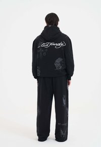 Person iført løs sort hoodie og bukser med hvid "Ed Hardy" logo og tatoveringslignende grafik, stående med ryggen til på en ensfarvet baggrund.