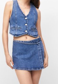 Jeanscropvest med V-ringning och fyra vita knappar; matchas med en matchande jeansminikjol med sidoknapp och sömdekor.