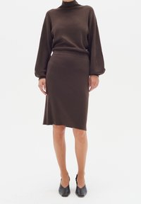Maglione marrone lavorato a maglia con collo alto e spalle scese, abbinato a una gonna fitted lunga fino al ginocchio nello stesso colore. Tacchi a punta neri.