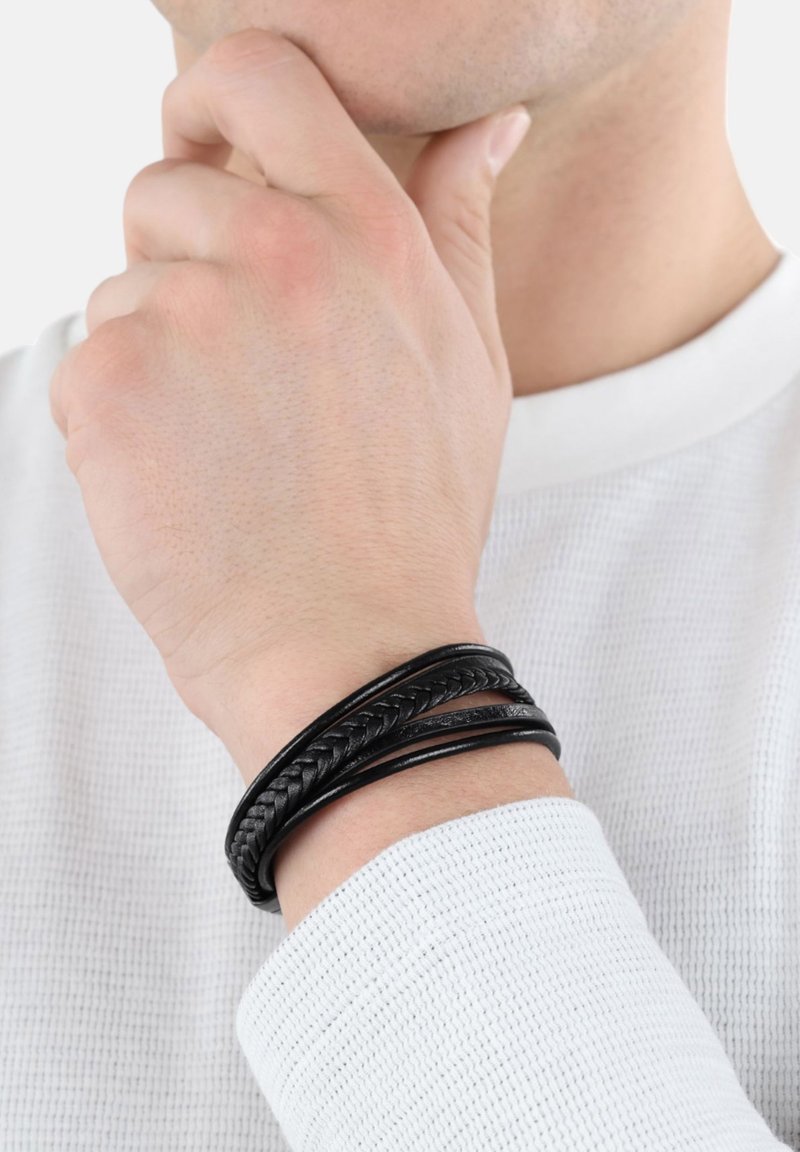 Bracciale in pelle nera composto da tre filamenti: uno liscio, uno intrecciato e uno testurizzato. Indossato su un polso con una camicia bianca e testurizzata.