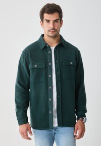 Maglione di pile verde con due tasche sul petto, chiusura a bottoni e colletto classico. Indossato sopra una maglietta bianca, con jeans a gamba dritta.