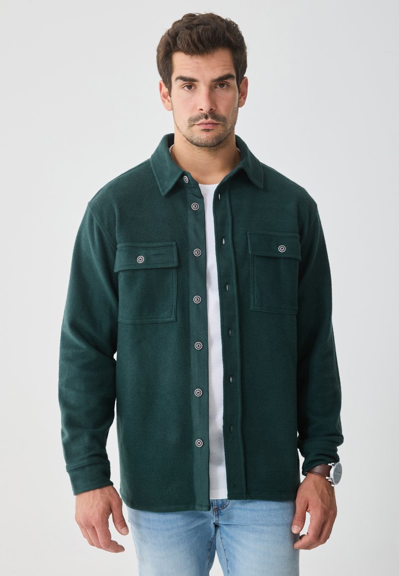 Maglione di pile verde con due tasche sul petto, chiusura a bottoni e colletto classico. Indossato sopra una maglietta bianca, con jeans a gamba dritta.
