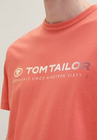 Korálové bavlněné tričko s kulatým výstřihem a krátkými rukávy. Na přední straně se nachází bílé tištěné logo "TOM TAILOR" a pod ním text v kontrastním designu.