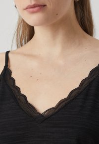 Μαύρο καμισόλ με V λαιμόκοψη που διαθέτει κυμματιστό δαντελένιο τελείωμα. Απαλό, υφασματοποιημένο ύφασμα με διακριτό μοτίβο, ρυθμιζόμενες τιράντες.