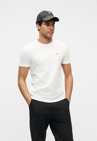 Άνδρας που φοράει λευκό κοντομάνικο μπλουζάκι με μικρό λογότυπο, μαύρο παντελόνι και σκουρόχρωμο τζιν καπέλο μπέιζμπολ, στέκεται με τα χέρια στις τσέπες.