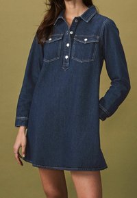 Džínové šaty s dlouhými rukávy, límečkem, dvěma předními kapsami a knoflíkovou légou. Tmavě modrá barva s mírně texturovanou tkaninou.