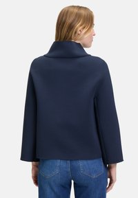 Marineblauer Pullover mit hohem Kragen, weiten Dreiviertelärmeln und glatter Textur; kombiniert mit hellblauen Jeans.