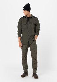 Camicia di flanella verde oliva con accenti neri sulle spalle, due tasche sul petto; abbinata a pantaloni cargo verde oliva e un berretto nero. Stivali marroni.