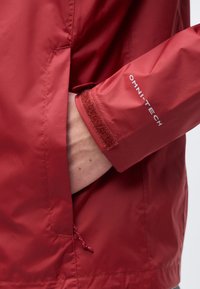 Main insérée dans la poche d'une veste rouge, manche avec une sangle Velcro ajustable étiquetée "OMNI-TECH" visible.