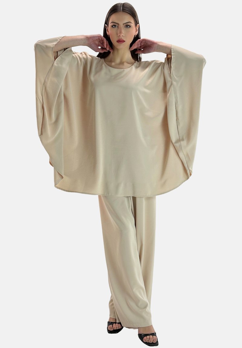 Top in satin beige con maniche ampie e orlo arrotondato, abbinato a pantaloni larghi coordinati. Texture liscia, minimo dettaglio.