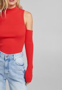 Röd ribbad ärmlös topp med hög krage och långa åtsittande ärmförlängningar, kombinerad med ljusblå högmidjade jeans.