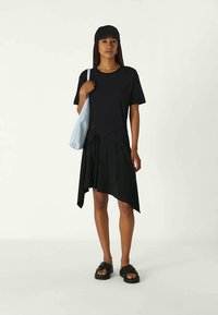 Mujer vistiendo un vestido negro asimétrico, una gorra negra, sandalias negras y llevando un bolso de hombro azul claro, de pie contra un fondo liso.