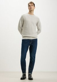 Maglione grigio chiaro con texture e scollo rotondo, abbinato a jeans blu scuro. Stivali neri alla caviglia completano l'outfit, posando su uno sfondo neutro.