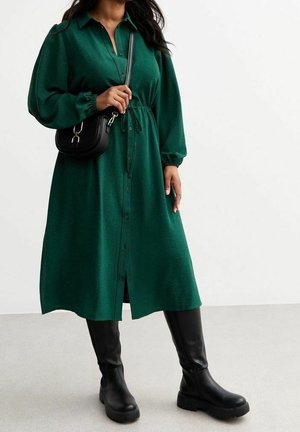 Vrouw draagt een lange-mouw midi-jurk in groen met knopen en een tailleband, een zwarte crossbodytas en zwarte kniehoge laarzen.