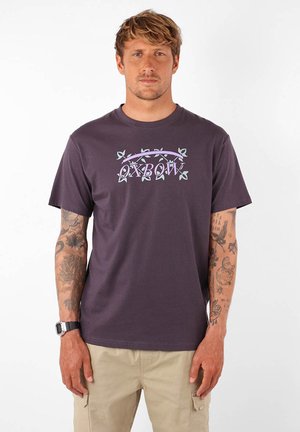 T-shirt en coton violet foncé avec un graphique "OXBOW" blanc et vert clair agrémenté de feuilles. Manches courtes, design col rond.