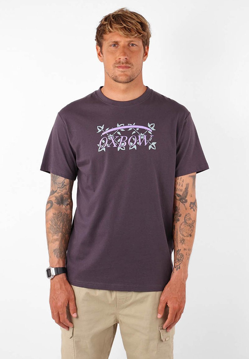 Camiseta de algodón de color púrpura oscuro con un gráfico en blanco y verde claro de "OXBOW" con acentos de hojas. Diseño de manga corta y cuello redondo.