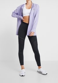 Felpa leggera zip-up viola, reggiseno sportivo bianco, leggings neri con dettagli in mesh e scarpe slip-on bianche con accenti neri.