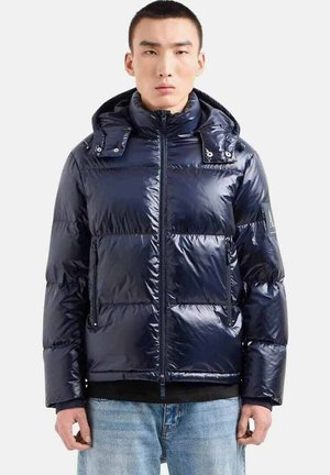 Giacca puffer blu navy con finitura lucida, cappuccio, chiusura con zip e bottoni a pressione. Presenta due tasche frontali e un branding discreto sulla manica.