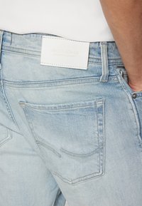Jeans de mezclilla azul claro con un aspecto desvaído, que presentan una etiqueta de cuero blanco con la marca "JACK & JONES" y dos bolsillos traseros con costuras curvas.